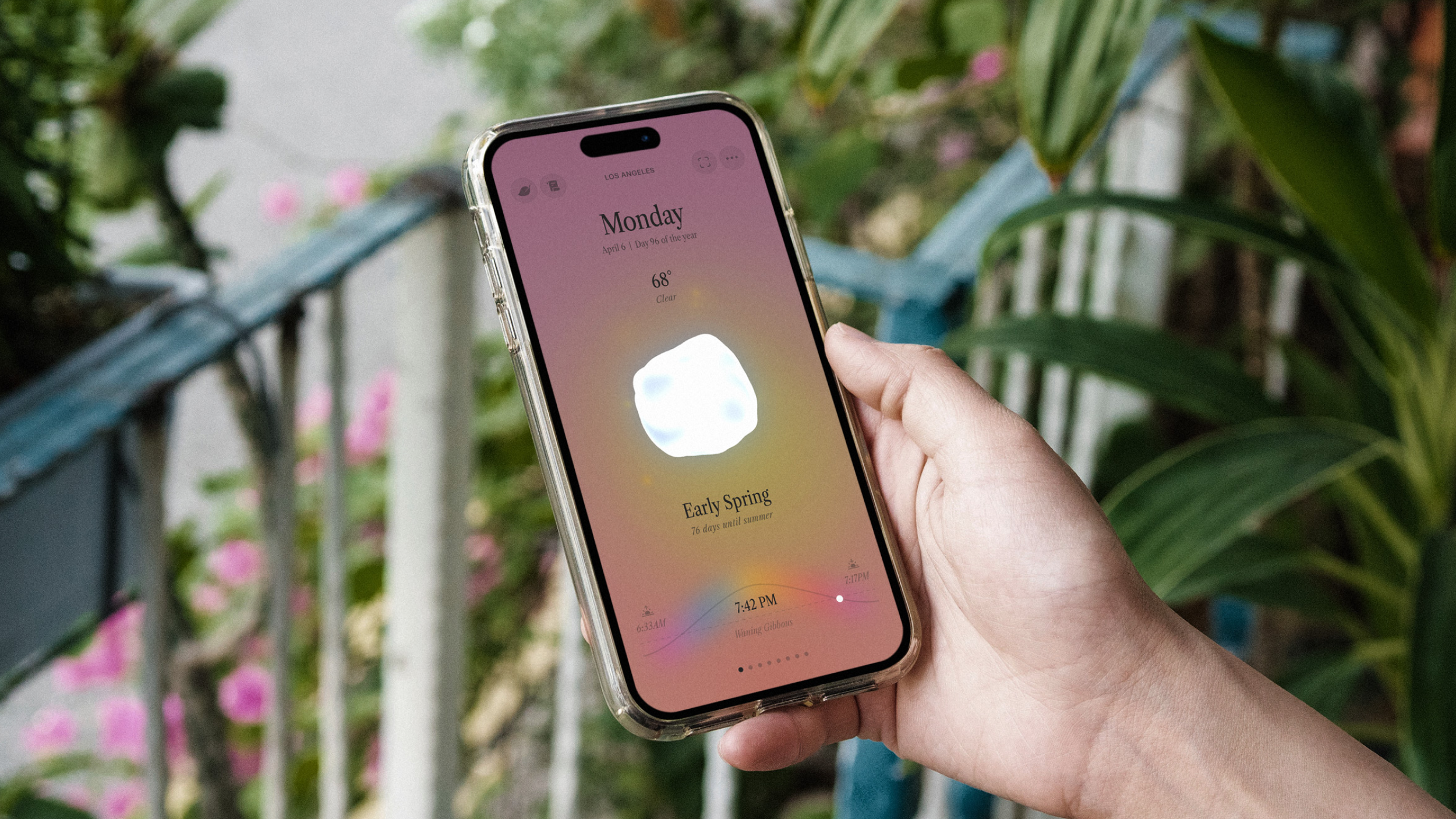 Hand holding an iPhone displaying a minimalist weather app with a pastel gradient background, showing Monday, 68 degrees, clear skies, and early spring timing.