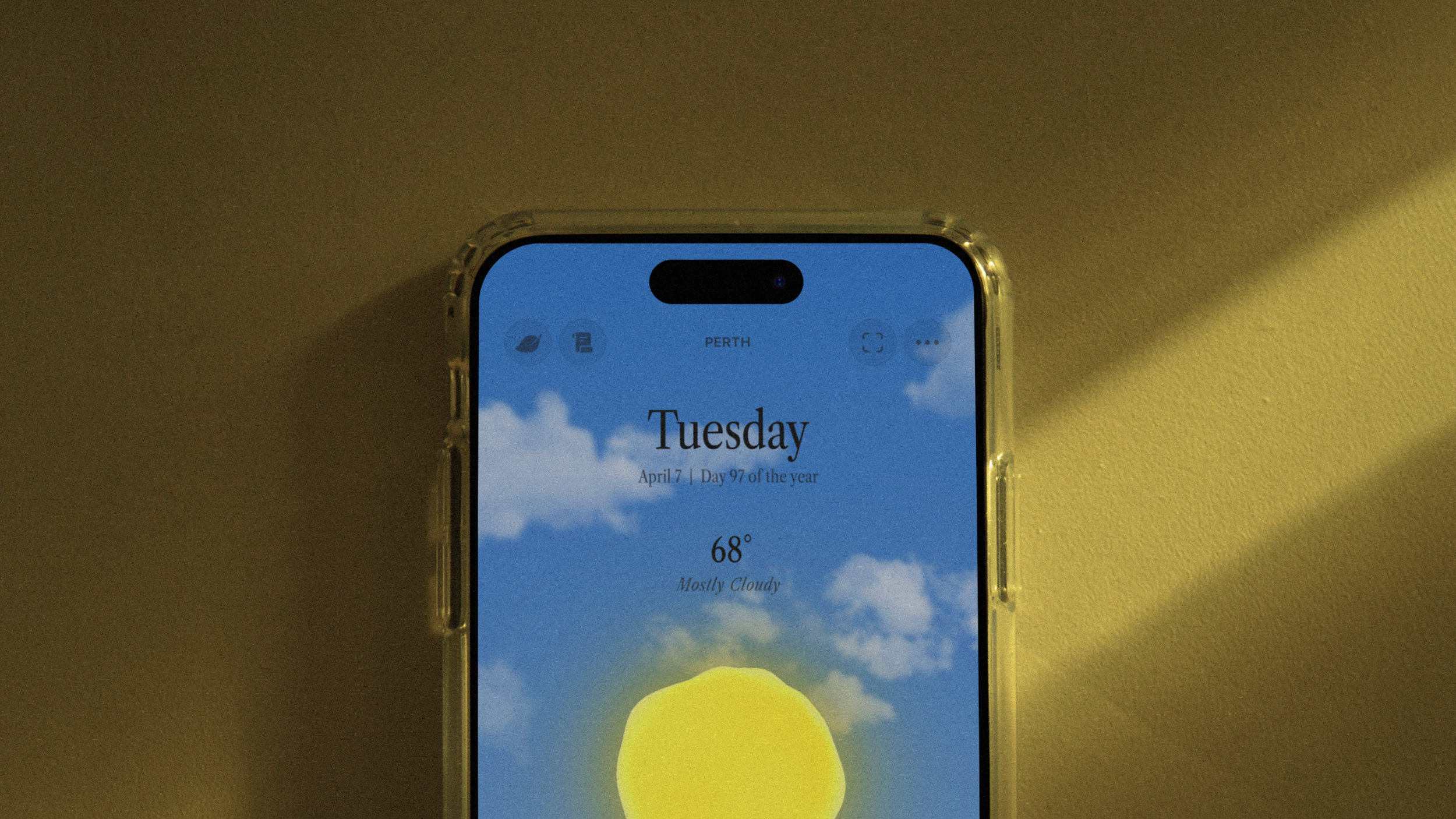 Close-up of an iPhone screen showing Ambient Almanac with blue sky and clouds, displaying Tuesday, 68 degrees, and mostly cloudy conditions.