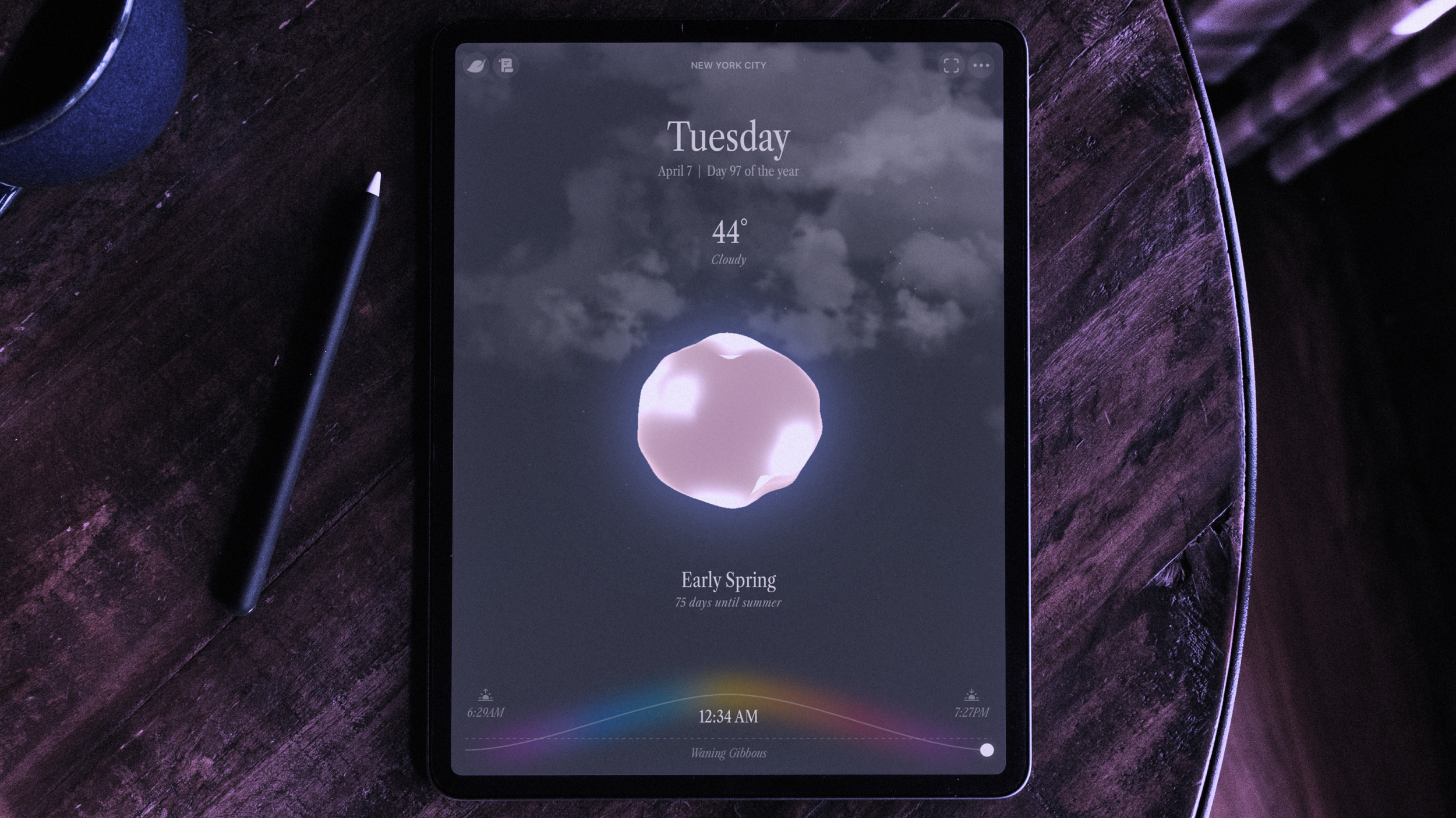 iPad on a wooden table displaying a dark-themed weather app interface with cloudy conditions, 44 degrees, and early spring information.
